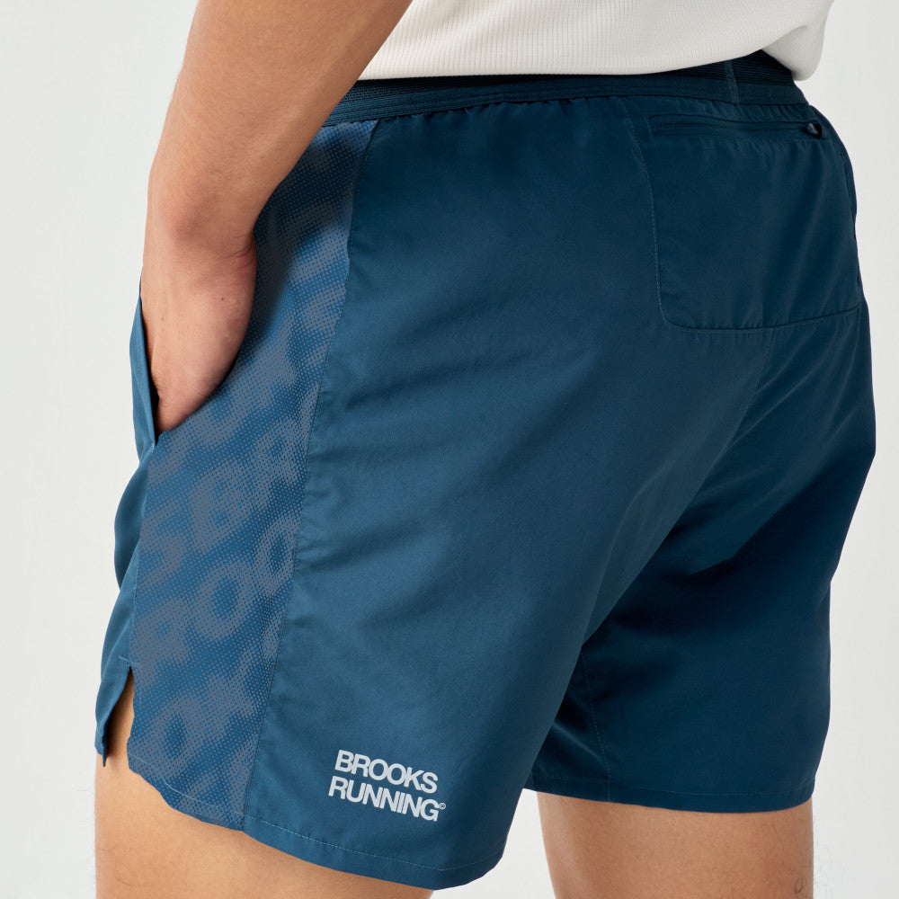 Close-up of a person wearing Brooks Running Dash shorts in dark teal blue, showing the side pocket, side slit, and white Brooks Running logo near the hem.