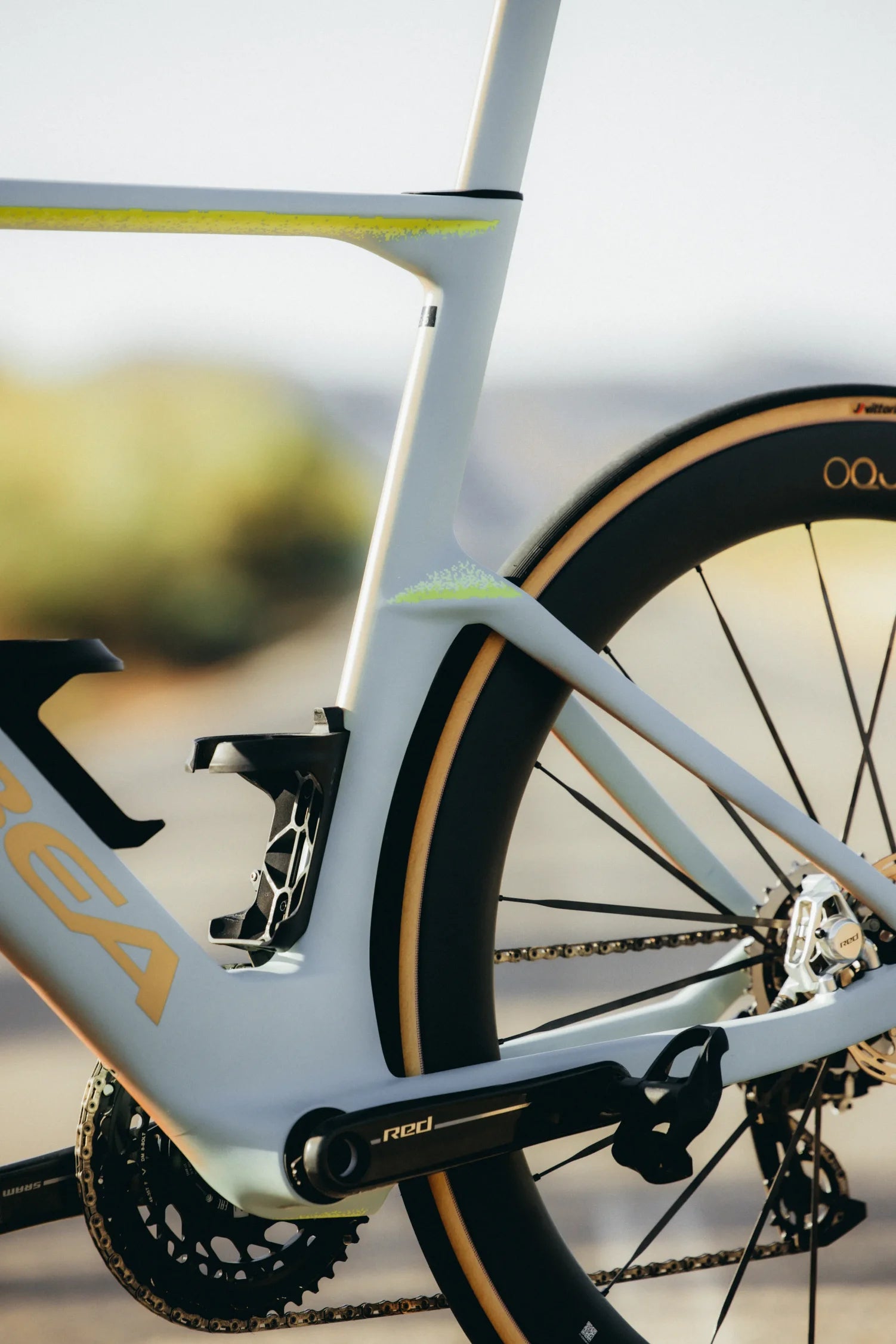 Close-up of an Orbea Aero M11e white and gold road bicycle frame with a black rear wheel, showing the seat tube junction, chainstay, crankset, and part of the tyre against a blurred outdoor background.