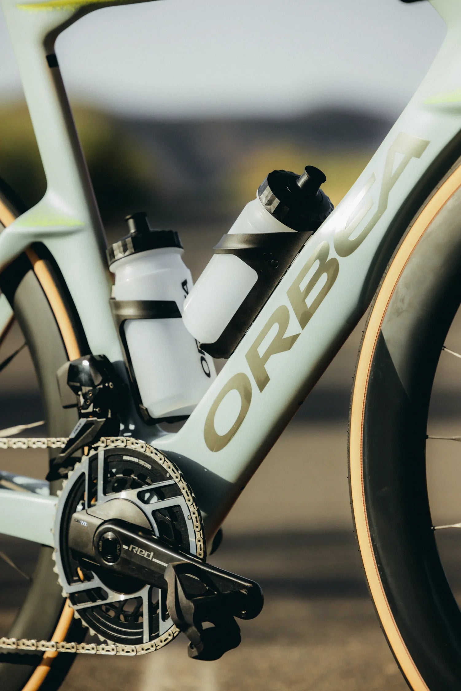 Close-up of an ORBEA Aero M11e road bicycle frame with two white water bottles mounted in the seat tube area, set against a blurred outdoor background.