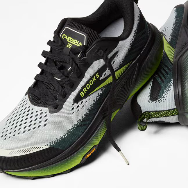 brooks cascadia 20 mens, road to trail gravel running and walking shoe, light grey and black with lime green details, one shoe sitting elevated ontop of a turned over one with untied laces