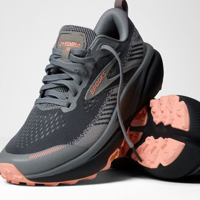 brooks cascadia 20 for women, road to trail gravel running and walking shoe, dark grey and black with peach detail, one shoe tipped over with one shoe sitting lifted on top with untied laces