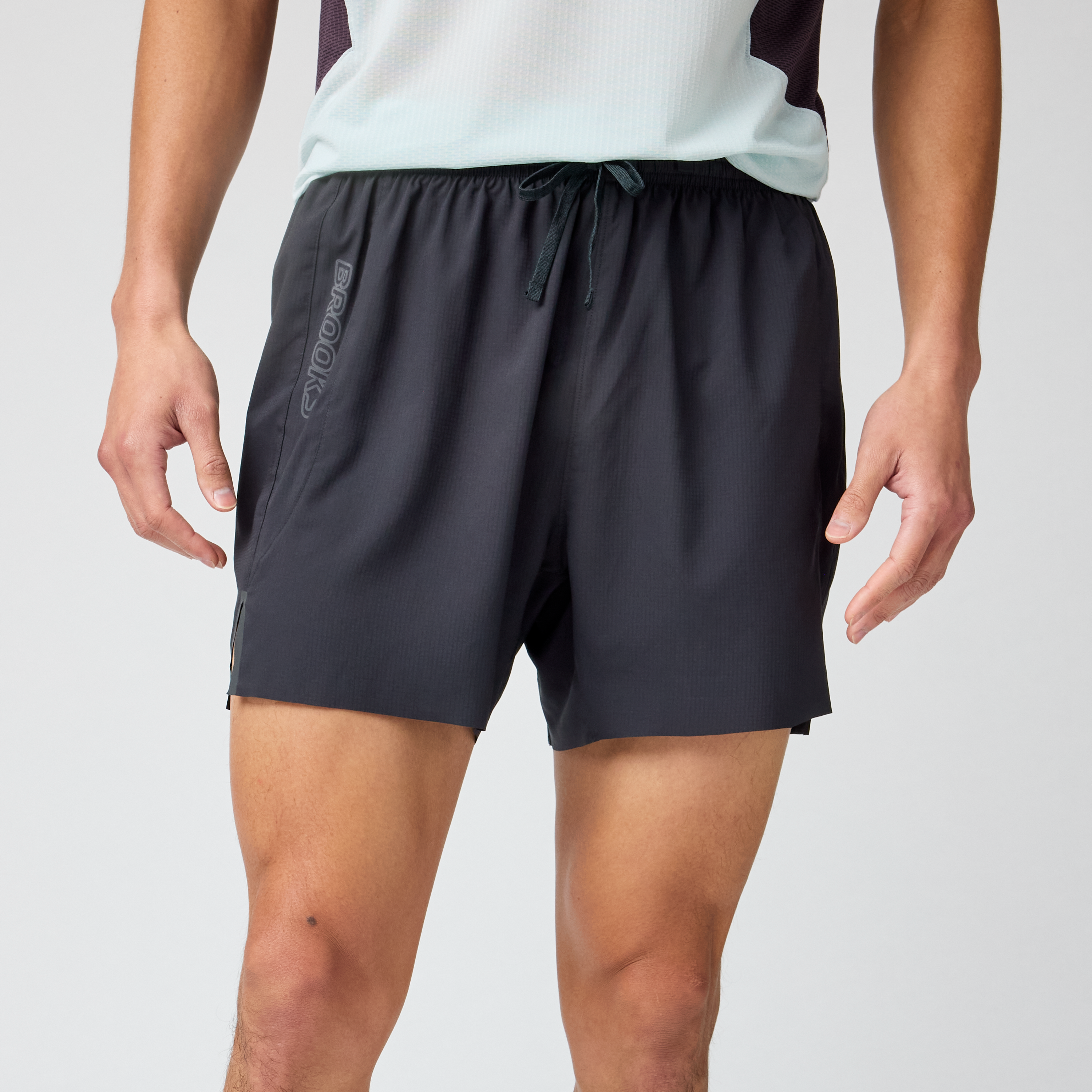 brooks cascadia short for men, lightweight running short for men with waist storage and zipper pockets, black colour, man wearing shorts with white top an dbackground