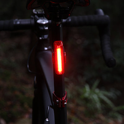 A close-up of a bicycle frame with a bright red Cateye rear light illuminated, photographed outdoors in a dark, blurred natural setting.