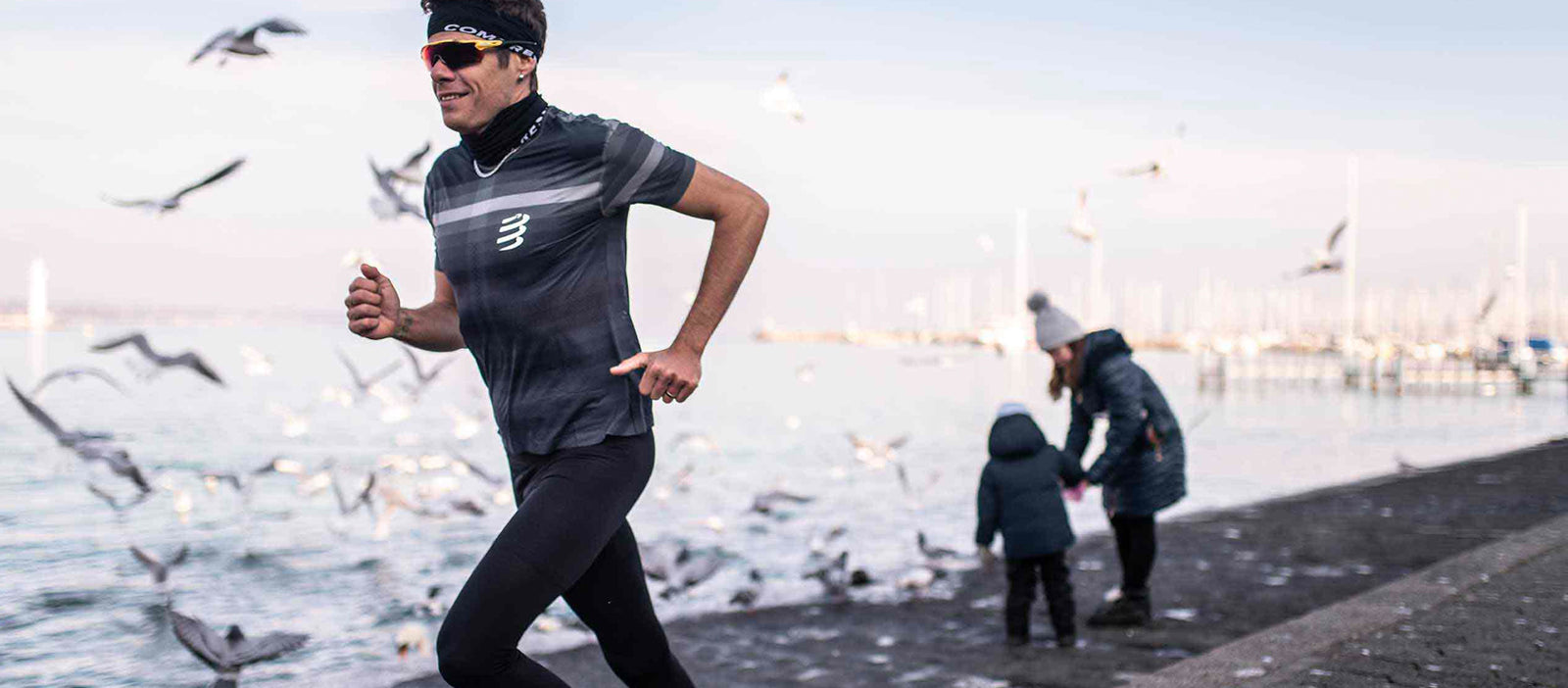 Person wearing compressport running on a path by a lake with children and seagulls in the background