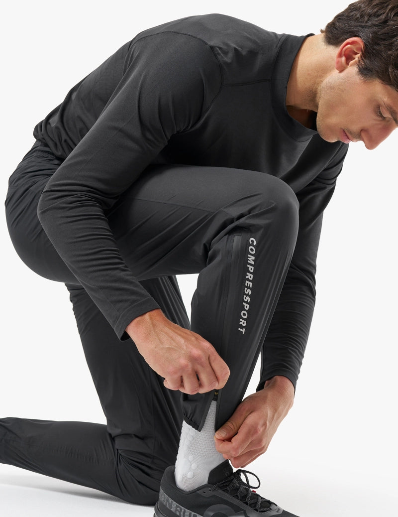 compressport hurricane pants 2.0, lightweight super breathable waterproof running pant, black colour, male model kneeling zipping ankle