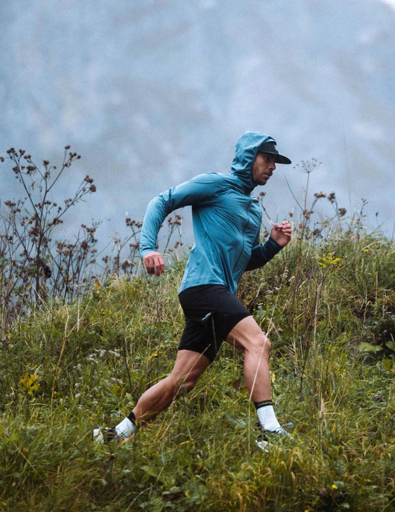 compressport tempest waterproof jacket, breathable and waterproof running jacket, deep aqua blue colour, man with hood up running in field