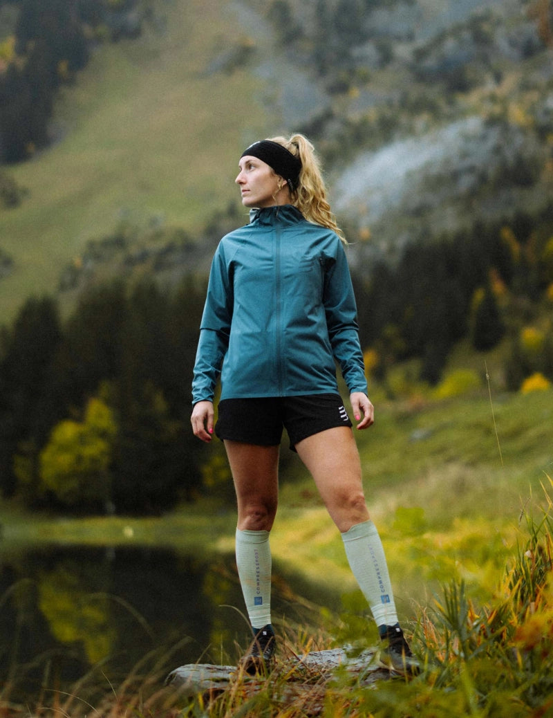 compressport tempest waterproof jacket for women, lightweight breathable and packable, dark aqua blue colour, model wearing jacket in mountains
