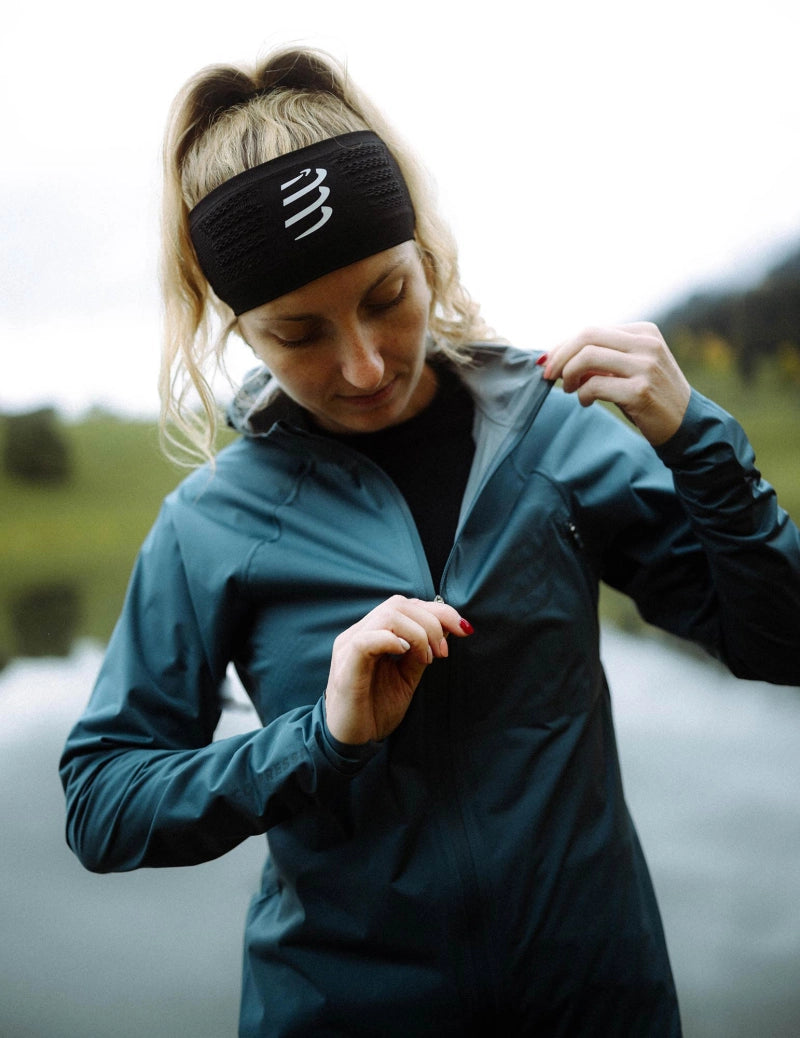 compressport tempest waterproof jacket for women, lightweight breathable and packable, dark aqua blue colour, model wearing jacket in mountains, up close model wearing jacket outside zipped open