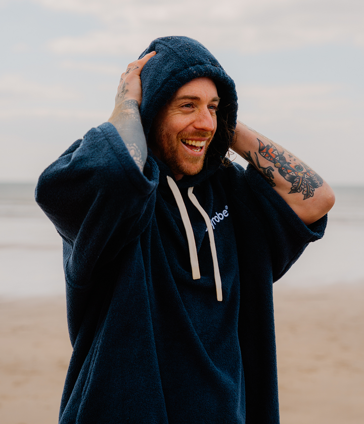 dryrobe organic cotton changing robe for swimming, soft and absorbent towelling robe for all gender, navy colour, man wearing dryrobe towel on a beach background