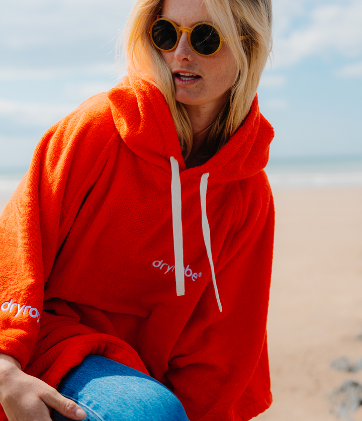 dryrobe organic cotton changing robe for swimming, soft and absorbent towelling robe for all gender, orange colour, woman wearing dryrobe towel on a beach background