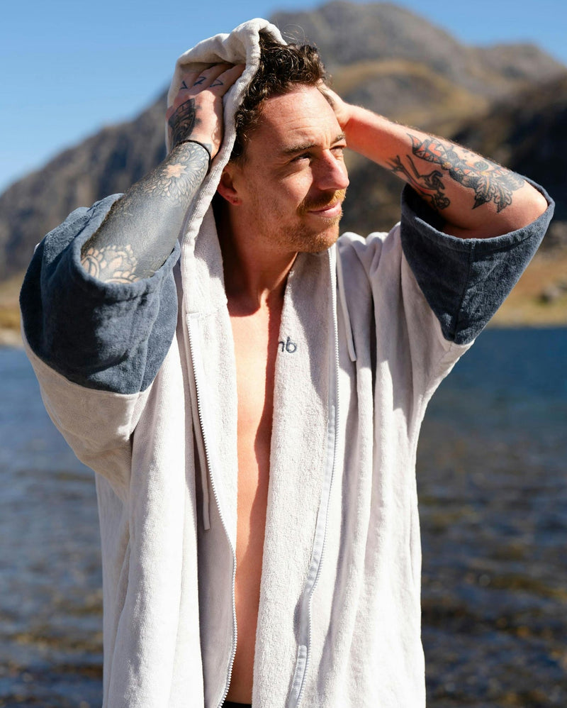 dryrobe towel robe for men and women, full zip towel robe for after swimming, light grey white colour with grey sleeve caps, man wearing dryobe outside drying off hair with hood 