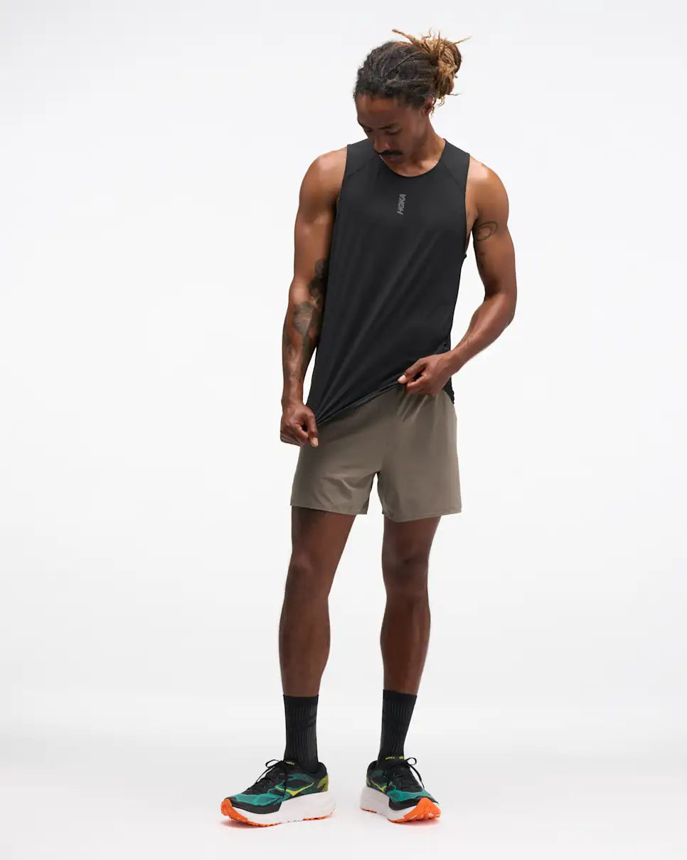 hoka everyrun tank top for men, light breathable running singlet , black colour with silver reflective HOKA logo, full body profile of a man wearing hoka tank with taupe shorts