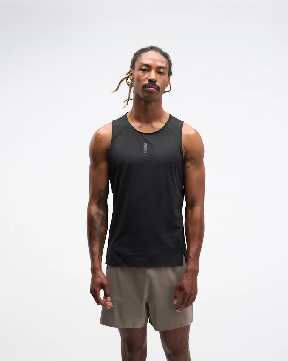 hoka everyrun tank top for men, light breathable running singlet , black colour with silver reflective HOKA logo, full body profile of a man wearing hoka tank with taupe shorts