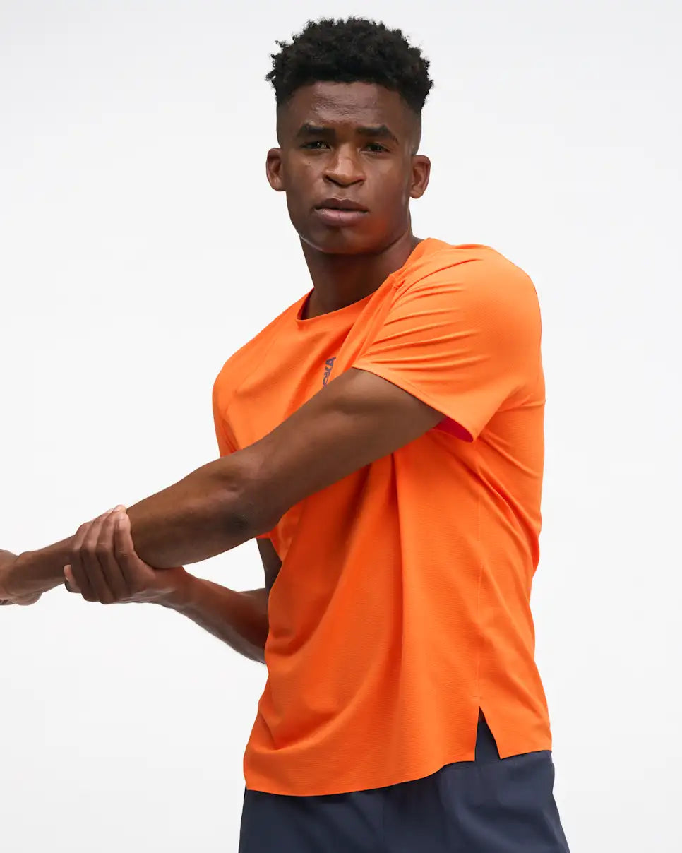 hoka everyrun short sleeve running t-shirt for men, bright orange colour with silver reflective HOKA logo, man wearing hoka tee with grey shorts on a white background