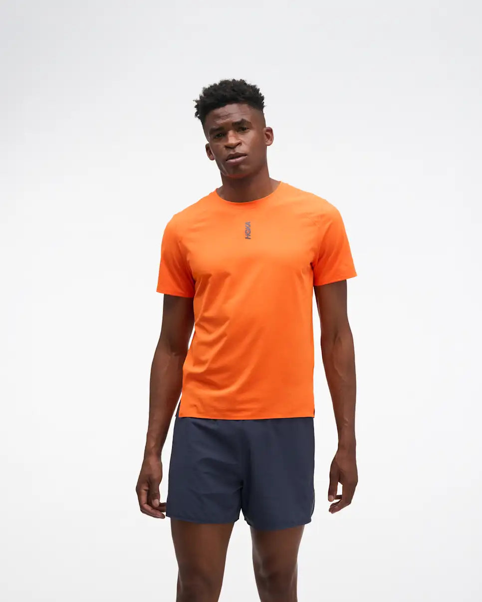 hoka everyrun short sleeve running t-shirt for men, bright orange colour with silver reflective HOKA logo, full body profile of a man wearing hoka tee with grey shorts on a white background