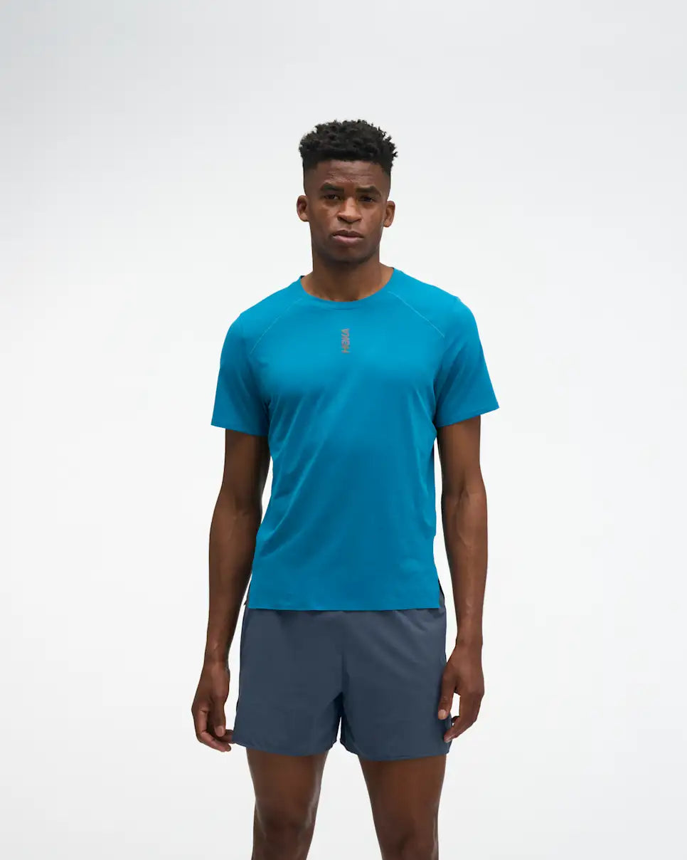 hoka everyrun short sleeve running t-shirt for men, blue colour with silver reflective HOKA logo, full body profile man wearing hoka tee with grey shorts on a white background