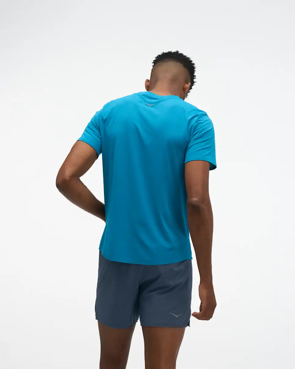hoka everyrun short sleeve running t-shirt for men, blue colour with silver reflective HOKA logo, back view of man wearing hoka tee with grey shorts on a white background