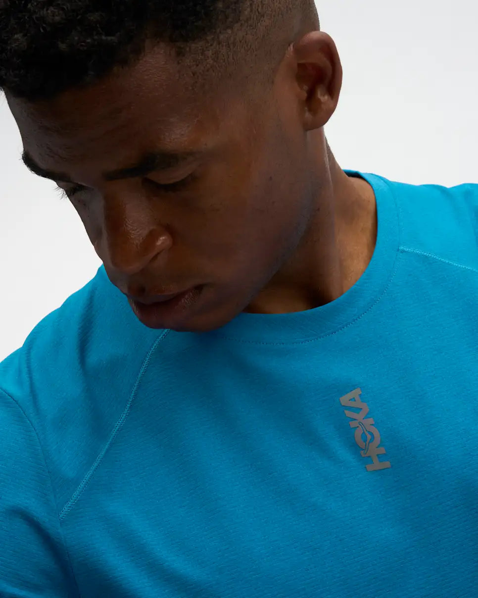 hoka everyrun short sleeve running t-shirt for men, blue colour with silver reflective HOKA logo, close up of a man wearing hoka tee with grey shorts on a white background