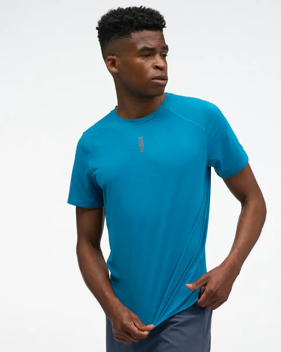 hoka everyrun short sleeve running t-shirt for men, blue colour with silver reflective HOKA logo, man wearing hoka tee with grey shorts on a white background