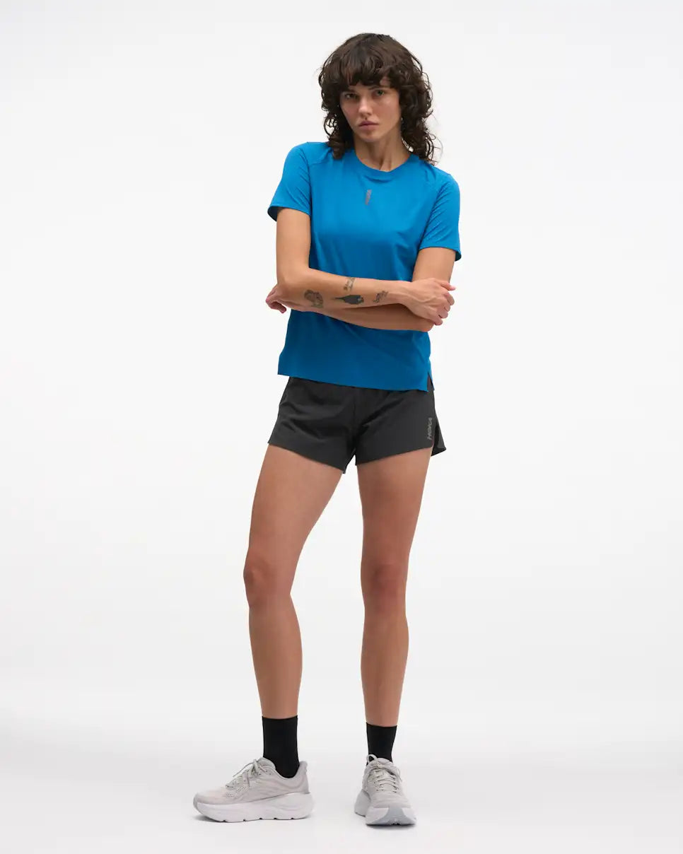 hoka everyrun short sleeve running t-shirt for women, blue colour with silver reflective logo, woman wearing hoka tee with black shorts on a white background