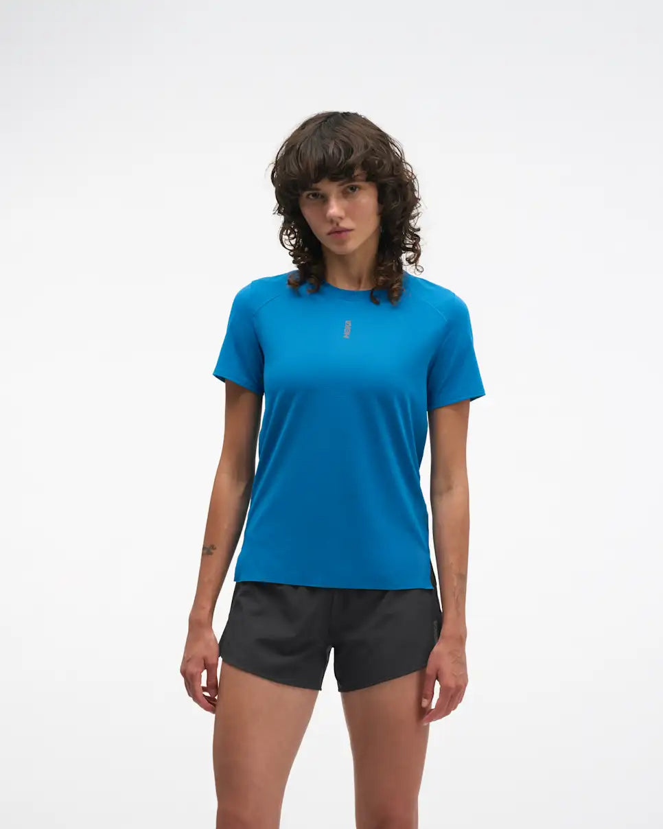 hoka everyrun short sleeve running t-shirt for women, blue colour with silver reflective logo, woman wearing hoka tee with black shorts on a white background