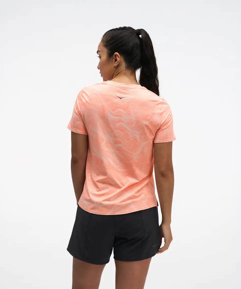 hoka everyrun short sleeve running t-shirt for women, peach colour with silver reflective logo, back view of woman wearing hoka tee with black shorts on a white background