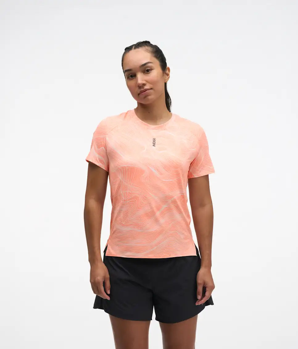 hoka everyrun short sleeve running t-shirt for women, peach colour with silver reflective logo, front view of woman wearing hoka tee with black shorts on a white background