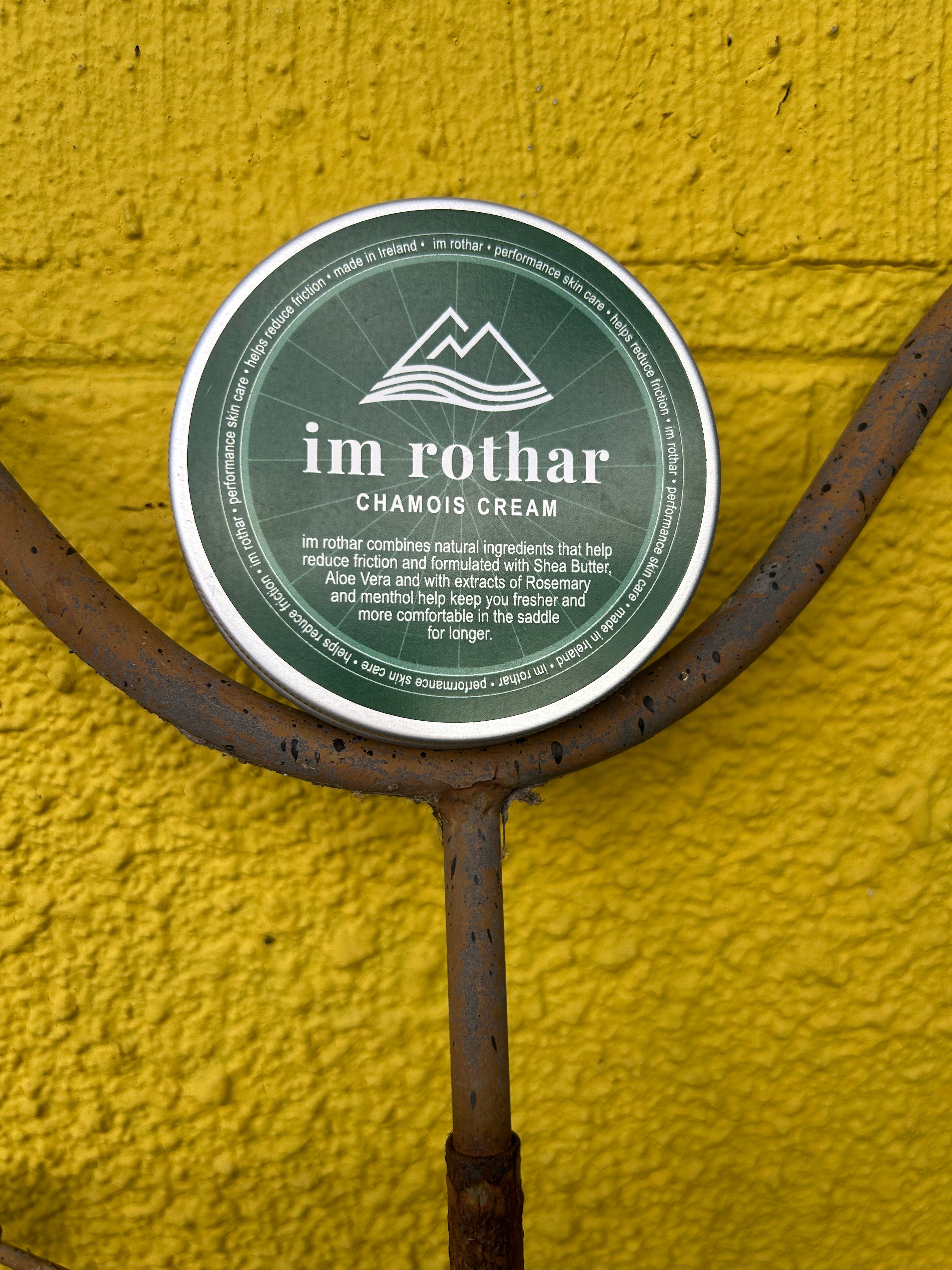 A round tin of im rothar Chamois Cream with a dark green label, mounted on a rusty metal bike stand against a bright yellow textured wall.