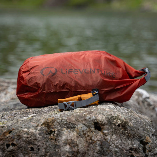lifeventure ultralight dry bag, 5 litre capacity, waterproof and durable storage bag, red rust colour, displayed on a rock with a body of water in the background