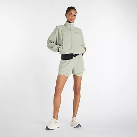 new balance athletics packable jacket for women, light wind breaker jacket that packs away into built in bag, light green colour, woman wearing nb jacket hands in pockets with matching shorts and white nb runners