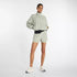 new balance athletics packable jacket for women, light wind breaker jacket that packs away into built in bag, light green colour, woman wearing nb jacket hands in pockets with matching shorts and white nb runners