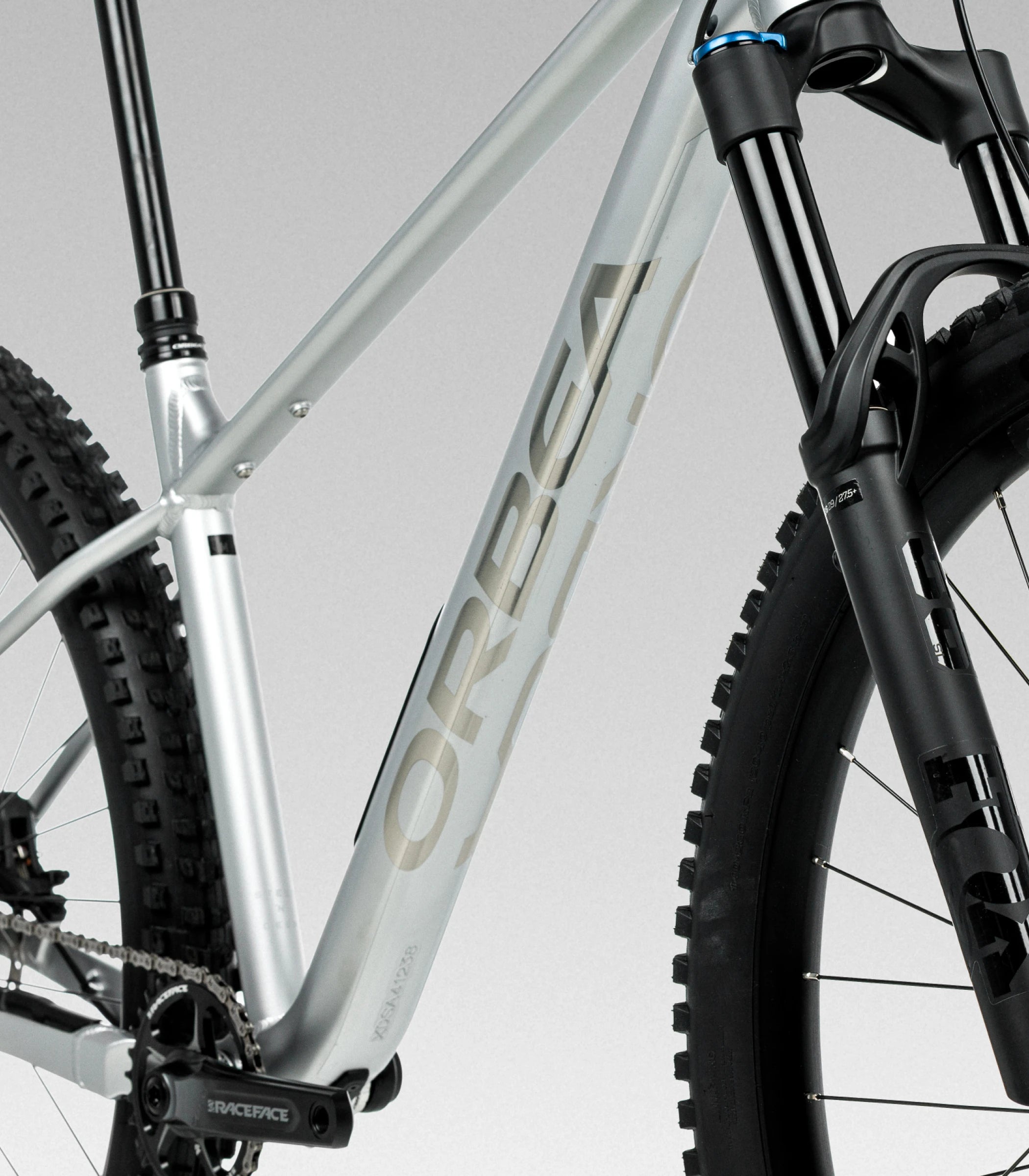 Close-up of an Orbea Laufey mountain bike frame and front suspension against a light gray studio background. Frame is metallic silver with large 'ORBEA' branding, and the front fork and tyres are black