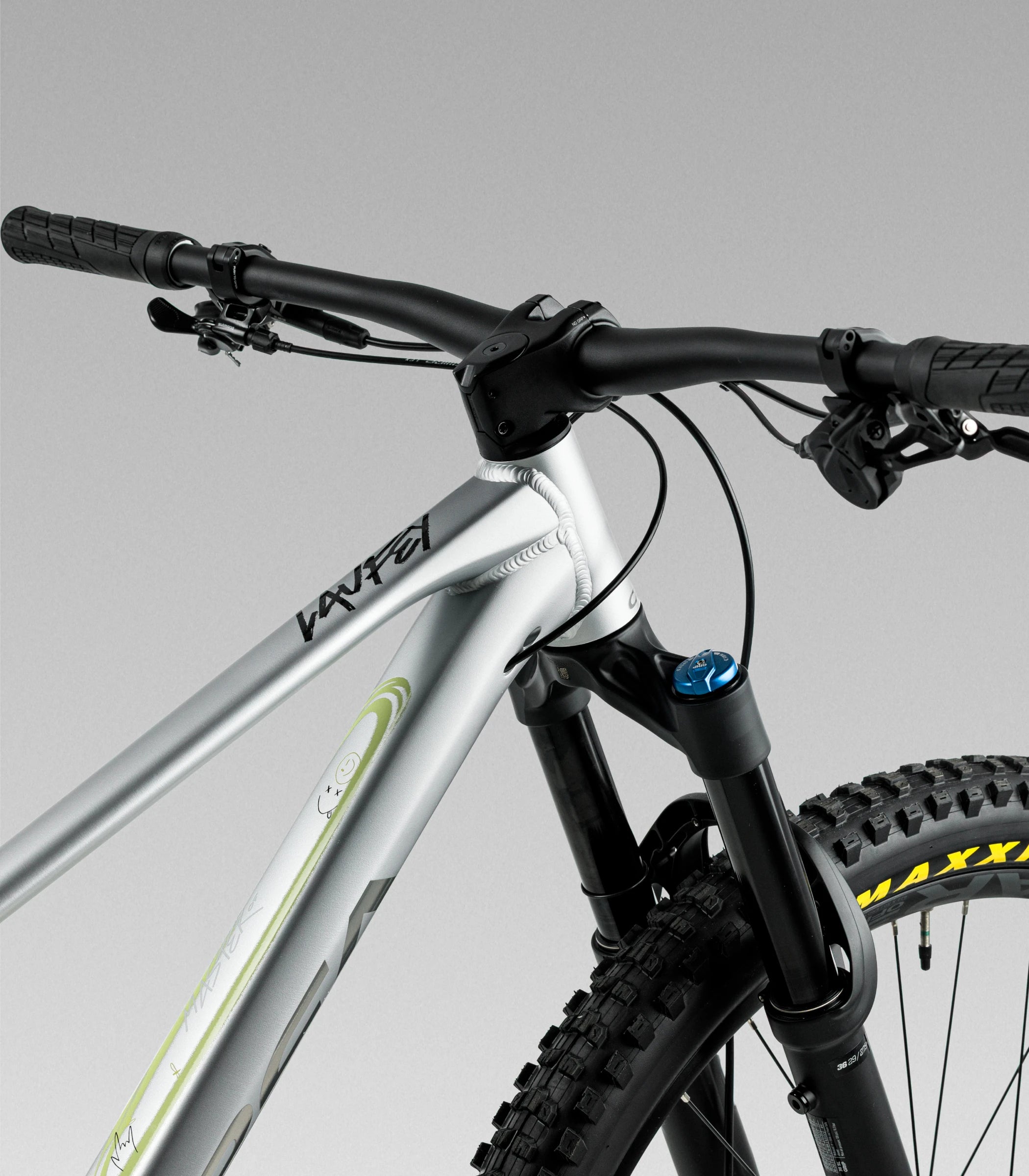 Close-up of a orbea laufey mountain bike frame and front fork on a gray studio background. The frame is metallic silver with black components, black handlebars, and a visible Maxxis tyre.