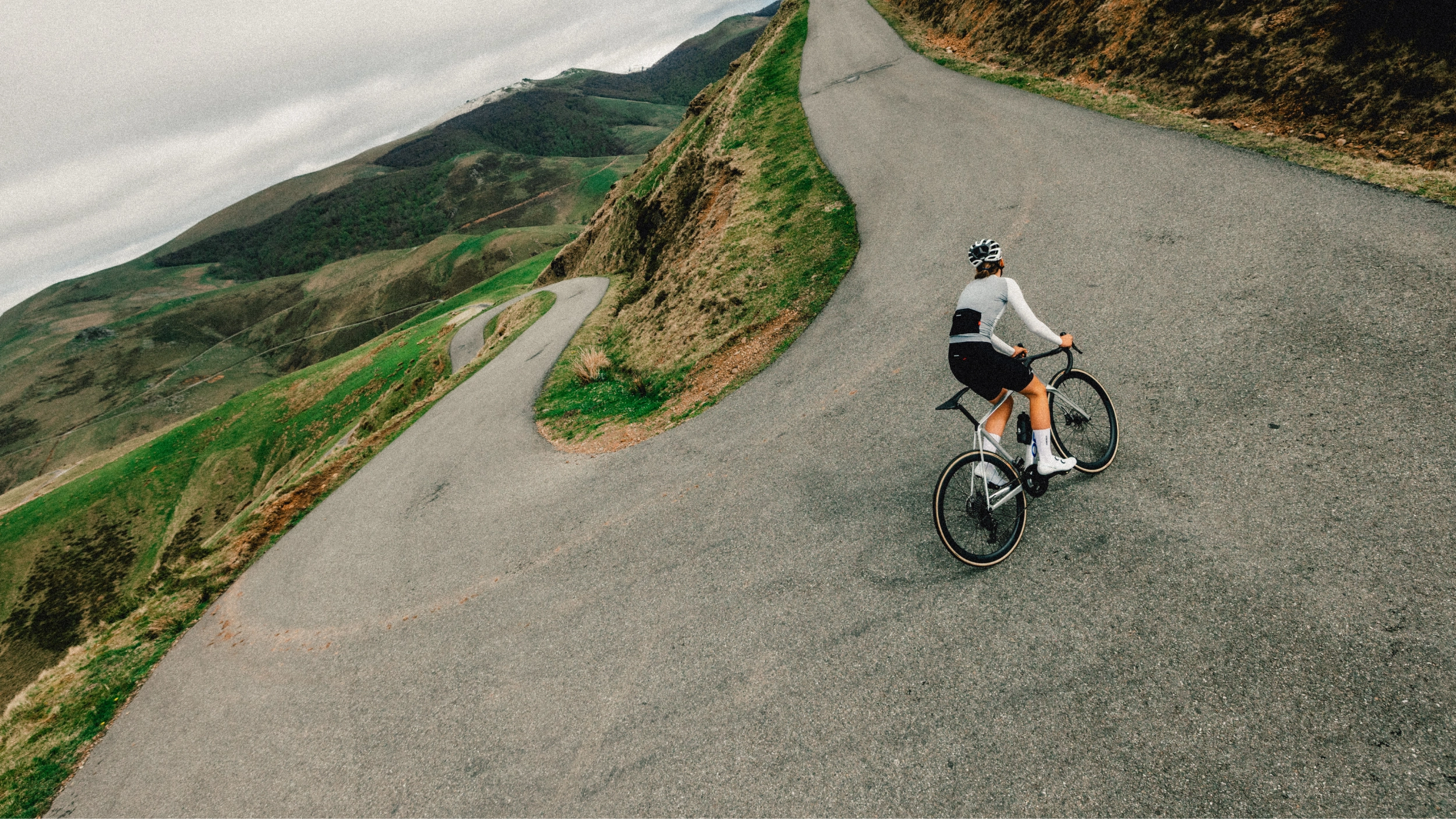 A cyclist rides an Orbea Orca road bike along a winding paved mountain road with grassy hills on both sides under an overcast sky.