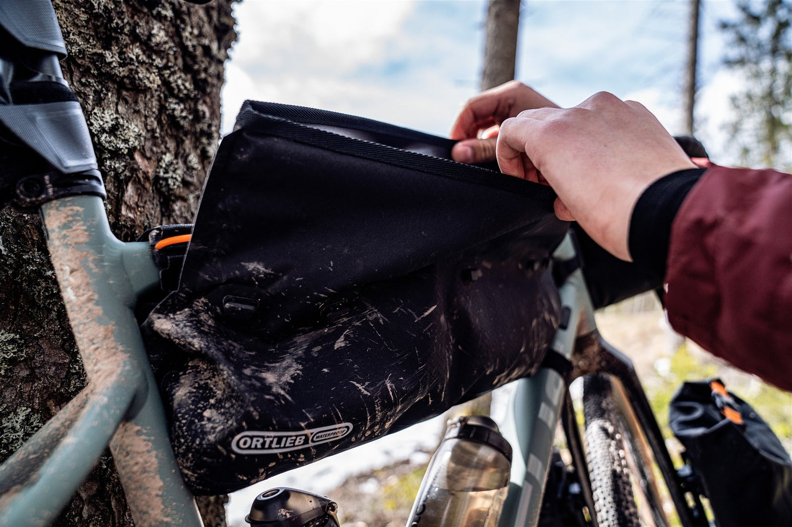Person adjusting a black Ortlieb bikepacking bag on a bicycle in a forest setting