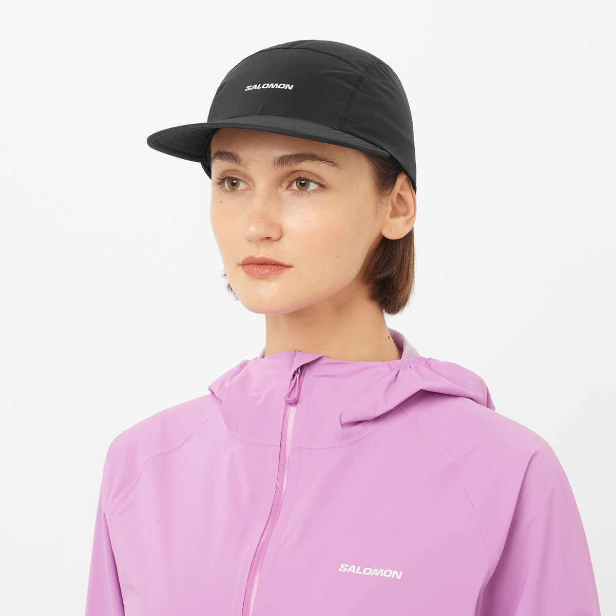 salomon bonatti pro cap with five panels, waterproof cap for running, black colour with a white logo, model wearing salomon cap with pink salomon jacket 