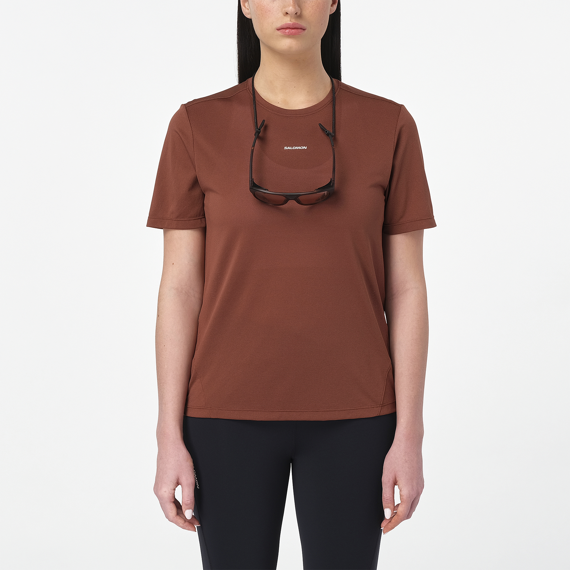 salomon shakeout core short sleeve running t-shirt for women, light and breathable run tee, dark burgundy colour with white salomon logo, model wearing salomon tee on white background with black leggings and sunglasses around her neck