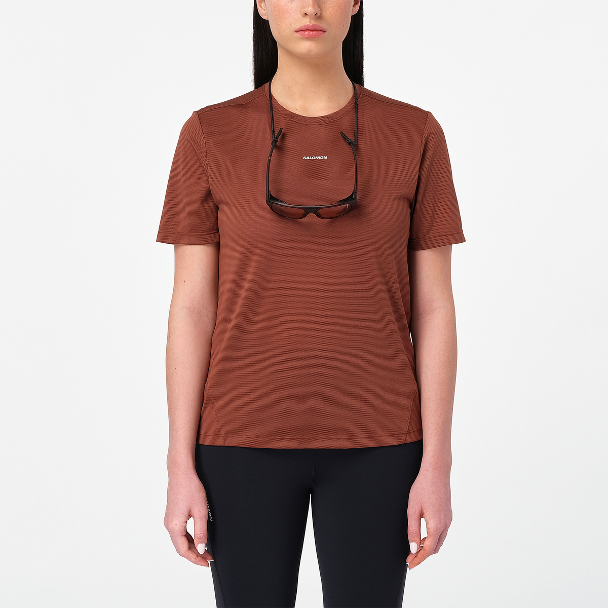 salomon shakeout core short sleeve running t-shirt for women, light and breathable run tee, dark burgundy colour with white salomon logo, model wearing salomon tee on white background with black leggings and sunglasses around her neck