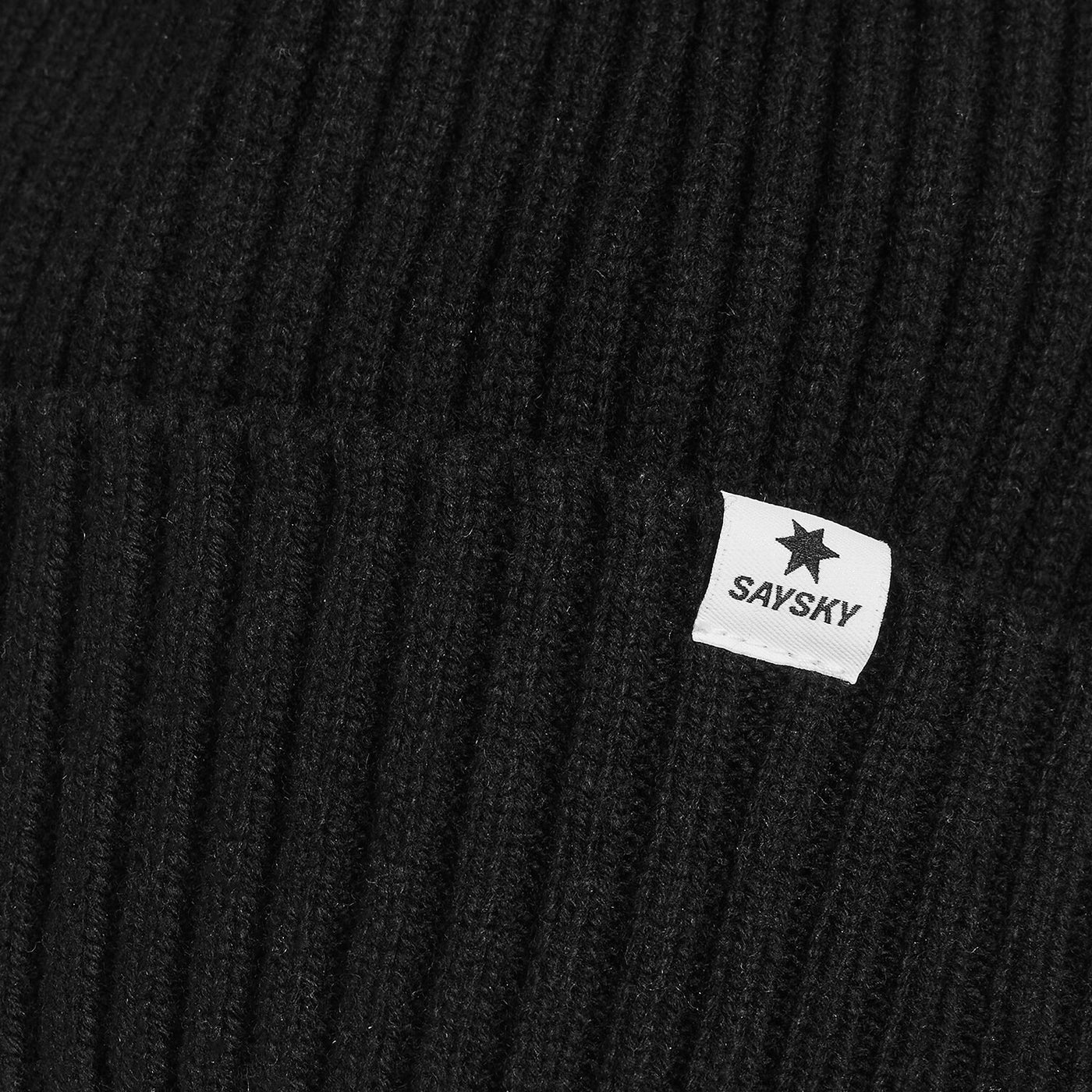 Close-up of a black ribbed knit beanie with a small white sewn label featuring a black star and the brand name SAYSKY.